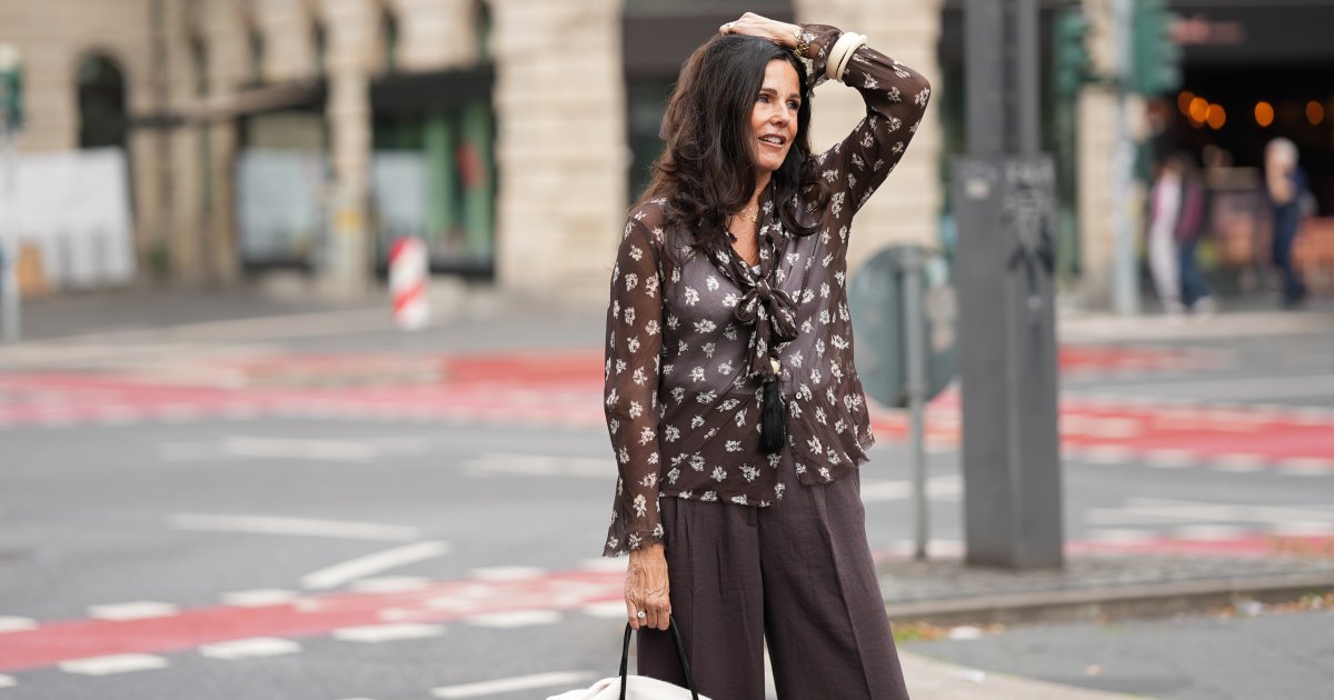 Angelika Hanel is seen wearing a chocolate brown flowing shirt with scarf collar, semi-sheer fabric, cream white floral print and button-up front from Massimo Dutti; a soft beige v-neck cashmere pullover with a relaxed silhouette and subtle ribbed cuffs and hem from Sa Su Phi; dark brown wide-leg linen blend trousers with a relaxed fit from H&M; a large black suede top-handle Soft Margaux 17 bag with softly structured sides, gold-tone buckles on the sides and a curved silhouette from The Row; a long black ceramic tassel cord necklace from Massimo Dutti; a golden necklace with a vintage Alhambra pendant in the shape of a cloverleaf from Van Cleef & Arpels; a golden Day-Date wristwatch from Rolex; a white gold pavé diamond tennis bracelet; an ecru colored Luca arm cuff set of two, made of synthetic resin from Sézane; a ring stacked with gold bands and a diamond from Marré; a diamond ring on July 27, 2025 in Frankfurt am Main, Germany.